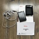 Na fotografii je černý přenosný MP3 přehrávač s displejem, bílá sluchátka a černé plastové pouzdro. Pod přístroji leží dvě krabice s červeným nápisem…