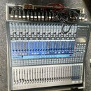 Míčovací pult PreSonus StudioLive 24.4.2 v šedé barvě s modrými sekcemi ovládání. Na horní hraně je připojeno množství kabelů a v pravém horním rohu…