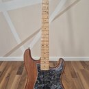 Elektrická kytara s hnědým dřevěným korpusem a světle hnědým javorovým hmatníkem. Na hlavě je viditelné logo Fender. Kytara má černý mramorovaný pick…