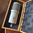 Kondenzátorový mikrofon stříbrné barvy v dřevěném kufru s černou pěnovou výplní. Na těle mikrofonu je nápis LAUTEN AUDIO, logo ve tvaru štítu s červe…