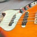 Elektrická basová kytara s korpusem v barevném přechodu od žluté přes oranžovou k červené. Nástroj má stříbrný plech ochranný (pickguard), dva černé…