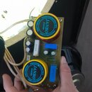 Fotografie znázorňuje elektronický obvod na zelené desce s dvěma velkými červenými válcovými součástkami. Obě součástky mají žlutý štítek s modrým kr…