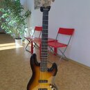 Elektrická basová kytara s pěti strunami a korpusem v barevném přechodu od žluté přes oranžovou k hnědé (sunburst). Na hlavě hmatníku je viditelný ná…