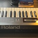 Černý digitální klavír s bílými a černými klávesami. Na přední hraně je velký nápis Roland a v pravém horním rohu logo Roland. Vlevo na ovládacím pan…