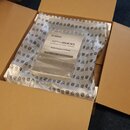 V otevřeném kartonovém boxu se nachází zařízení zabalené v průhledném plastu a obložené ochrannou fólií s černými symboly. Na zařízení je viditelný n…