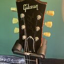 Fotografie zachycuje hlavu černé elektrické kytary s bílým logem Gibson a nápisem Les Paul MODEL. Nástroj má šest ladicích kol v krémové barvě a je u…