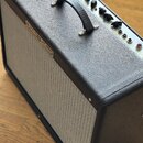 Kytarový kombozesilovač s černým texturovaným povrchem a světle šedou textilní maskou. Na předním panelu je nápis „Fender Deluxe“. Horní ovládací pan…