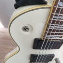 Detail bílé elektrické kytary s hnědým hmatníkem a dvěma černými snímači. Na spodní části těla je částečně viditelný nápis GRIND. Povrch instrumentu…