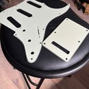 Dvě bílé plastové krycí desky (pickguardy) pro elektrickou kytaru položené na černém židli. Jedna deska má výřezy pro tři snímače a ovládací prvky, d…