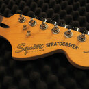 Fotografie zachycuje hlavu elektrické kytary z jasného dřeva s šesti stříbrnými ladičkami. Na povrchu je černý nápis Squier BY FENDER® a STRATOCASTER…