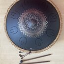 Černý kovový tongue drum s pleteným okrajem v béžové barvě. Uprostřed je vyrytý ornament mandaly a ve spodní části nápis „Sonic Energy“ s logem. K ná…
