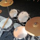 Fotografie zobrazuje bicí sadu s černými korpusy a kovovým hardwarem. Na činelech je vidět značka PAISTE. Součástí sady jsou dva tom-tomy, snare drum…