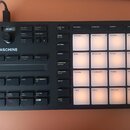 Černý kontrolér Maschine s mřížkou 16 podsvícených padů. Na levé straně je bílý nápis MASCHINE, rotační ovladač a řada tlačítek s popisky jako VOLUME…