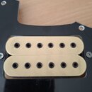 Fotografie zachycuje elektrický snímač typu humbucker s kovovými kryty v zlaté barvě. Snímač je osazen do černého plastového pickguardu a je připevně…