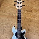 Bílý basový ukulele v držáku na dřevěné podlaze. Na hlavě hmatníku je nápis KALA a pod ním UBASS. Nástroj má tmavý hmatník, černý pickguard a čtyři č…
