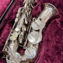 Stříbrný saxofon v otevřeném kufru s fialovou výstelkou. Na zvonu je čitelně napsáno „ASS“, „SUPER“, „model Replica“ a „MADE IN“. Povrch nástroje vyk…