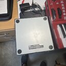 Fotografie zachycuje spodní stranu stříbrného, čtvercového elektronického zařízení s gumovými nožičkami v rozích. Na spodní straně je nalepený štítek…