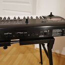 Černý digitální klavír umístěný na černém stojanu. Na horní části je bílé logo Roland a nápis JUNO-C. Na zadním panelu jsou viditelné popisky konekto…