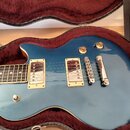Modrá elektrická kytara s tvarem singlecut uložená v tvrdém kufru s červeným polstrováním. Nástroj má dva humbuckery se zlatými rámečky, dva ovládací…