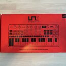 Červená krabice s obrázkem syntetizátoru. Na krabici je nápis SYNTHPRO a v dolní části text PARAPHONIC DUAL FILTER ANALOG SYNTHESIZER. V levém horním…