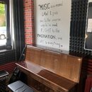 Hnědé pianino s vysokým leskem a doprovodnou černou polstrovanou stolečkou. Na nástroji nejsou vidět žádné značky ani loga. Povrch pianina je hladký…