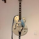 Bílá polodupatá elektrická kytara s modrým pickguardem a chromovaným hardwarem. Na hlavě hmatníku je nápis GRETSCH a na pickguardu je svisle umístěný…