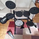 Elektronická bicí souprava s černým rámem a černými pady. Na modulu, na snare padu i na pedálu je vidět bílý nápis Roland. Souprava obsahuje pět padů…