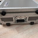 Šedý transportní kufr s hliníkovými hranami a kovovými rohy. Na horní části je nalepený štítek s nápisem ROCKBOARD. V dolní části jsou dvě černé kole…