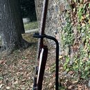 Tmavě hnědá elektrická basa s hmatníkem z tmavého dřeva, umístěná v černém kovovém stojanu. Nástroj je zobrazen z profilu, nejsou vidět žádná loga an…