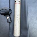 Stříbrný kondenzátorový mikrofon s kovovou síťkou a červeným kroužkem v horní části. Povrch těla mikrofonu vykazuje výrazné opotřebení, šmouhy a stop…