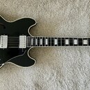 Černá polodupatá elektrická kytara s bílým lemováním a hnědými f-otvory. Na hlavě je viditelné logo Gibson. Nástroj má dva humbuckery, tři ovládací k…