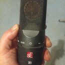 Černý kondenzátorový mikrofon s kovovou síťkou. Na těle je červené logo SE a pod ním bílý nápis X1. V horní části jsou viditelné značky -10 dB a symb…