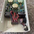 Fotografie zachycuje vnitřek bílého kovového šasi s elektronickým plošnicovým spojem. Na desce jsou vidět tři zlaté válcové kondenzátory, z nichž jed…