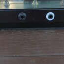 Detailní pohled na černý panel s dvěma konektory. V pozadí jsou vidět krabice s nápisy BOSS PS-5 SUPER Shifter a BOSS GEB-7 Bass Equalizer. Nad panel…