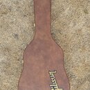 Hnědý tvrdý kufýr na akustickou kytaru s texturovaným povrchem. V dolní části je umístěno žluté logo Gibson. Kufýr leží na suché trávě.
