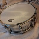 Stříbrný kovový snare drum s bílými blanami. Na horní blaně je černý oválný štítek s nápisy SONOR, White Medium a v dolní části menším písmem MADE IN…