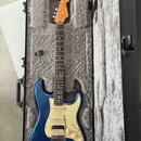 Modrá elektrická kytara typu Stratocaster s bílým pickguardem a javorovým hmatníkem v otevřeném černém tvrdém kufru s šedým polstrováním. Na hlavě ná…