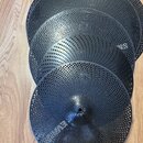 Čtyři černé perforované elektronické činely různých velikostí v kovovém provedení. Jsou rozloženy přes sebe na dřevěné podlaze. Na nejmenším činele v…