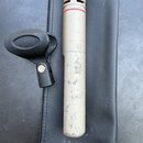Kondenzátorový mikrofon s kovovým těllem v béžové nebo zlatavé barvě a stříbrnou mřížkou. Povrch těla vykazuje výrazné opotřebení a odřeniny, v horní…