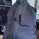 Černý polotvrdý obal na basovou kytaru s několika zipovými kapsami a popruhymi. Obal stojí na černém stojanu v interiéru. Povrch materiálu je jednotn…