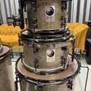 Tři bubny v barvě stříbrného glitru s chromovaným hardwarem. Na dvou horních bubnech je vidět čtvercový bílý štítek s logem a nápisem Jelen DRUMS. Po…