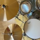 Sada bubnů a činelů v modrošedé barvě. Na blanách jsou viditelné nápisy EVANS a REMO. Činely mají nápis paiste pst. Povrch nástrojů vykazuje běžné zn…