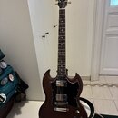 Tmavně hnědá elektrická kytara s dvojsečným tvaru těla a černým pickguardem. Na hlavě hmatníku je zlaté logo Gibson. Nástroj má dva humbuckery, stříb…