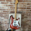 Elektrická kytara typu Stratocaster v červeně hnědé barvě s bílým pickguardem a světle hnědým hnedobým hmatníkem. Na hlavě je viditelné logo Fender S…