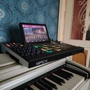 Černý sampler MPC s integrovaným dotykovým displejem, na kterém je zobrazeno rozhraní softwaru. Na přední hraně přístroje je bílý nápis MPCX. Zařízen…