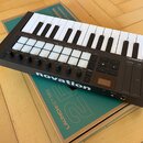 Černý MIDI kontrolér s klaviaturou, 16 bílými pady a několika ovládacími knoflíky. Na spodní hraně je bílý nápis novation a v pravém horním rohu ozna…