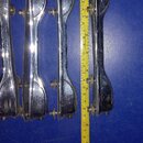 Čtyři chromované kovové držáky, pravděpodobně součásti mechanismu bicích. Vedle nich je přiložen žlutýv metr, který ukazuje délku cca 20 cm. Povrch k…