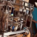 Detailní pohled na chromovaný snare drum značky Pearl. Na korpusu je viditelný černý obdélníkový štítek s bílým logem Pearl a nápisem JAPAN. Povrch n…