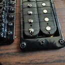 Detailní pohled na černý humbucker snímač a část černého mostu elektrické kytary. Snímač je osazen čtyřmi viditelnými šrouby a čtyřmi polosférickými…