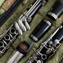 Černý klarinet s kovovými klapy uložený v zeleném polstrovaném pouzdře. Nástroj je rozložen na několik částí, včetně stříbrného zounu. Na horním díle…